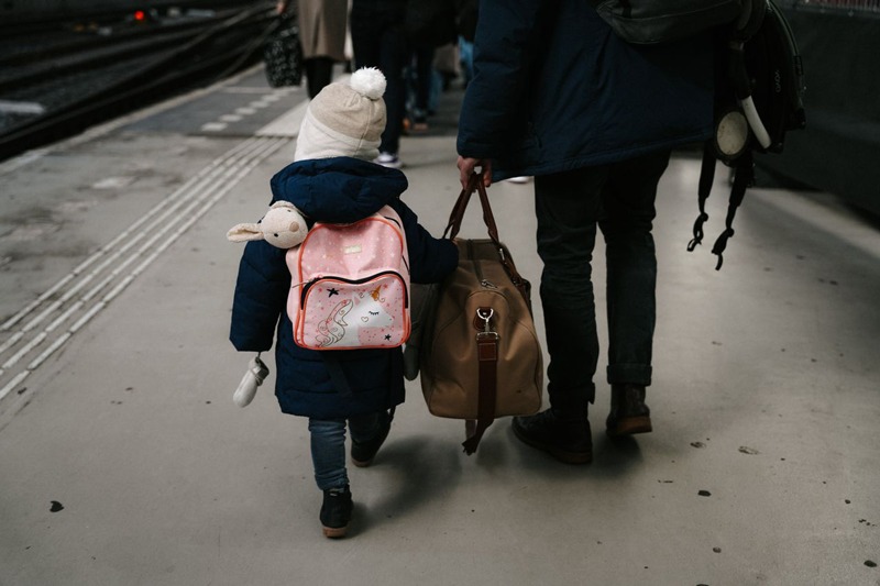 A child with a backpack and suitcase being walked through a train station